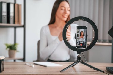 Kürsüdeki akıllı telefon kayıt yapıyor. Çevrimiçi ana sınıf. Beyaz elbiseli genç ve güzel bayan ofiste..