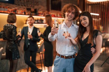 Kadın ve erkek ayakta duruyor ve poz veriyor. Güzel giyinmiş bir grup insan içeride yeni yılı kutluyorlar..