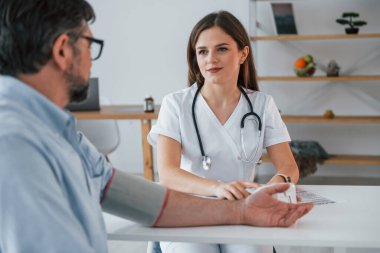 Kadın doktor adamın kan basıncını ölçüyor. Ofiste beyaz önlüklü profesyonel bir sağlık görevlisi var..