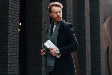 Elinde gazete tutuyordu. Sakallı şık adam binanın yanında..