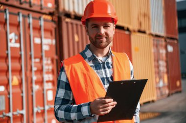 Elinde tabletle ayakta duruyor. Erkek işçi konteynırlarla birlikte olay yerinde..