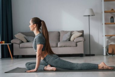 Bacakları esnetiyorum. İnce vücutlu ve yoga elbiseli genç bir kadın evde..