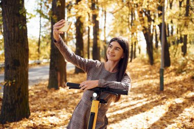 Genç kadın elektrikli scooter 'ın üzerinde dururken akıllı telefon kullanarak selfie çekiyor..