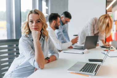 Olgun bir kadın dizüstü bilgisayarla masada oturuyor. Profesyonel iş adamlarından oluşan bir grup ofiste..