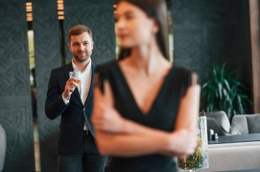 Siyah takım elbiseli ve elinde içki olan bir adam elbiseli bir kadına bakıyor..