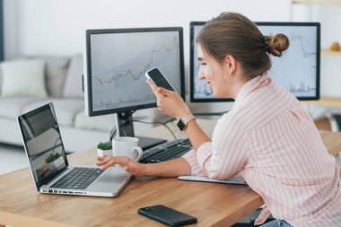 Akıllı telefon kullanıyor. Günlük giysiler içinde kadın borsacı ofiste bilgisayar başında çalışıyor..