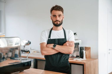 Kollarını kavuşturmuş duruyorsun. Beyaz gömlekli ve siyah önlüklü kafe çalışanı içeride..