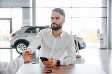 Masanın yanında oturuyor. Beyaz elbiseli genç adam araba galerisinde..