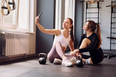 Oturup selfie çekiyoruz. Spor elbiseli iki kadın spor salonunda birlikte spor yapıyorlar..