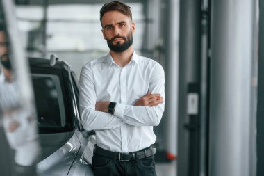 Kollarını kavuşturmuş duruyorsun. Beyaz elbiseli genç adam araba galerisinde..