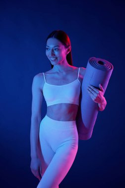 Egzersiz için hazır. Yoga minderiyle. Spor elbiseli genç bayan stüdyoda neon ışıklarıyla bekliyor..