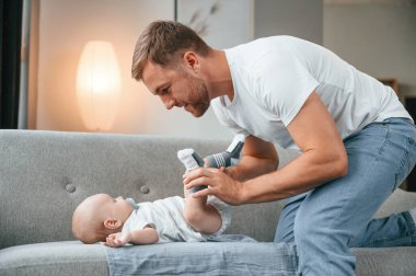 Bebek kanepede uzanıyor. Bebekli baba evde, oğluyla ilgileniyor..