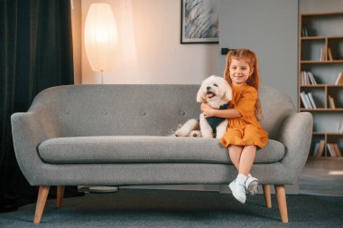 Sarı elbiseli küçük kız kanepede oturuyor ve evde köpeğiyle oturuyor..