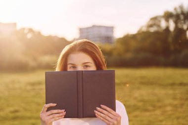 Kitabı tutuyor ve kameraya bakıyor. Genç kız güneşli bir günde sahada güzel bir hafta sonu geçiriyor..