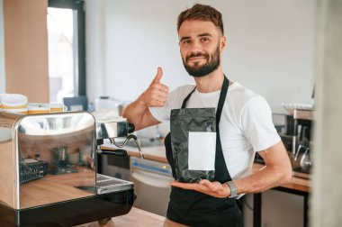 Elinde çiğ kahve paketi var ve parmağını kaldırıyor. Beyaz gömlekli ve siyah önlüklü kafe çalışanı içeride..