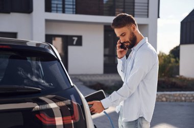 Aracı şarj etmek ve telefon kullanmak. Genç şık adam, gündüz vakti elektrikli arabayla geziyor..