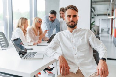 Masanın yanında oturan genç adamın portresi. Profesyonel iş adamlarından oluşan bir grup ofiste..