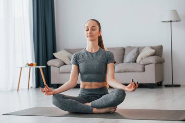 Lotus pozu. İnce vücutlu ve yoga elbiseli genç bir kadın evde..