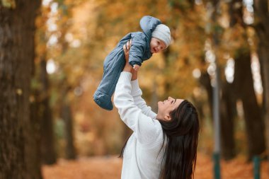 Güzel anne küçük oğlunu elinde tutuyor. Sonbahar ormanında dikilirken.