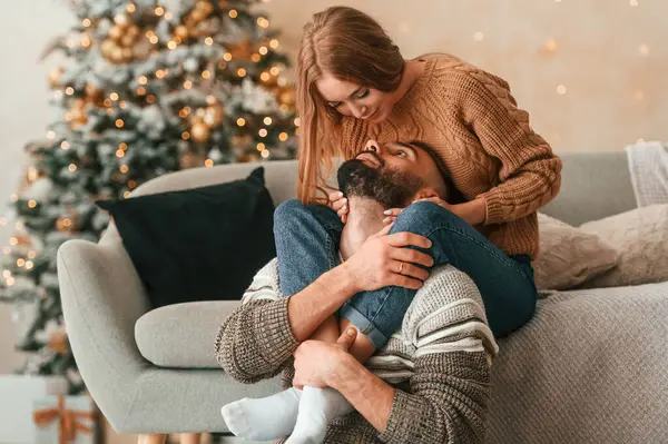 Kadın kanepede, erkeğini bacaklarından kucaklıyor. Güzel genç çift yeni yılı evde kutluyor..