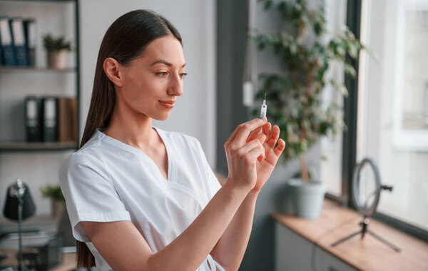 Holding syringe. Young beautiful female doctor in white coat is in the office.
