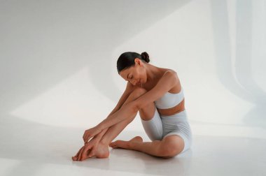 Rahatlamış, yerde oturuyor. İnce vücutlu, beyaz bir kadın stüdyoda..