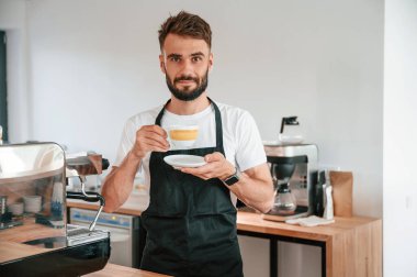 Ayakta ve bardağı tutarken. Beyaz gömlekli ve siyah önlüklü kafe çalışanı içeride..