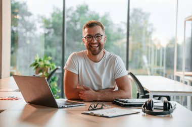 Oturuyor ve gülümsüyor. Modern ofiste resmi giyinmiş olumlu bir adam çalışıyor..