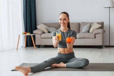 Portakal suyu ve meyve suyuyla. İnce vücutlu ve yoga elbiseli genç bir kadın evde..