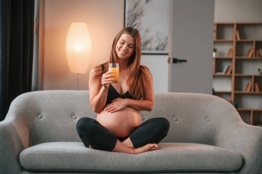 Elinde portakal suyuyla. Kanepede. Güzel hamile kadın evde..