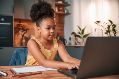 Çizim yapmak için dizüstü bilgisayar ve kağıtla masada oturuyordum. Gündüz vakti evde rahat giyinen sevimli siyah kız var..