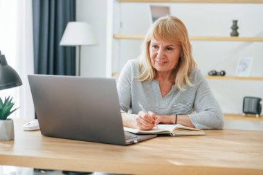 Masada dizüstü bilgisayarla oturuyor. Kızıl saçlı yaşlı kadın evde yalnız..