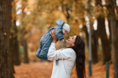 Güzel anne küçük oğlunu elinde tutuyor. Sonbahar ormanında dikilirken.