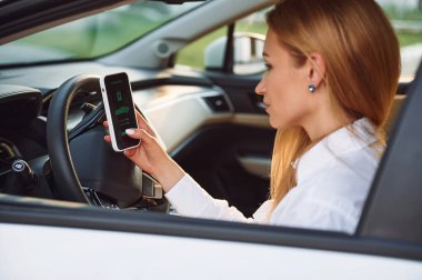 Akıllı telefonla içeride oturmak. Beyaz elbiseli genç bir kadın gündüz vakti elektrikli arabasıyla geliyor..