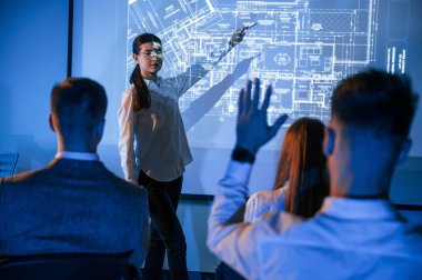 İşçilerle konuşan kadın lider, borsa şirketinin projektörüyle ilgili planı gösteriyor..