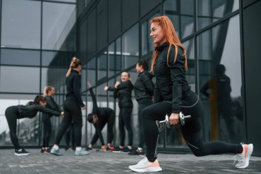 Dambıllarla çömelmek. Bir grup sportif kadın dışarıda, siyah binanın yakınında..