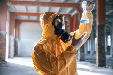 Radyoaktif maddelerle dolu bir test tüpü. Kıyamet sonrası binanın harabelerinde kimyasal koruma kıyafeti giymiş bir adam..