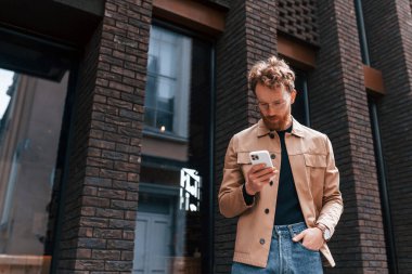 Akıllı telefon kullanarak internet bağlantısı. Sakallı, haki renkli ceketli ve kot pantolonlu şık bir adam binanın yanında..