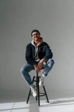 Şapkalı ve gözlüklü manken bir sandalyede oturuyor. Yakışıklı adam stüdyoda beyaz arka plana karşı..