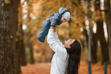 Güzel anne küçük oğlunu elinde tutuyor. Sonbahar ormanında dikilirken.