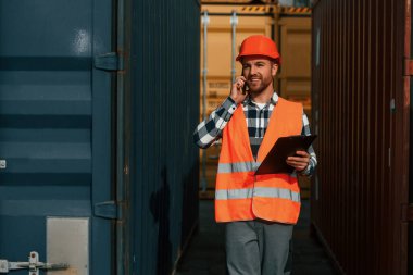 Telefon kullanarak ve tablet tutarak. Erkek işçi konteynırlarla birlikte olay yerinde..
