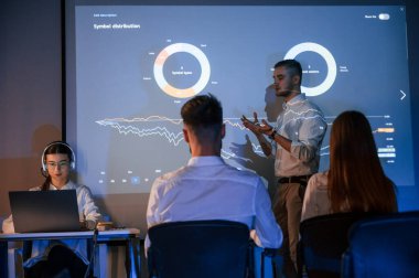 İnsanlar oturuyor. Erkek lider çalışanlarla konuşuyor. Borsa şirketinin projektörüyle ilgili planı gösteriyor..