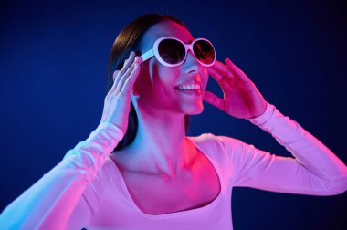 Gülümsüyor ve güneş gözlüğü takıyor. İçerideki neon ışıkta duran genç bir kadının portresi..