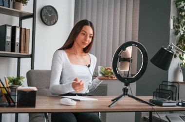 Beslenme uzmanı kamerada salata gösteriyor. Beyaz elbiseli genç ve güzel bayan ofiste..