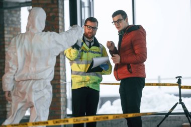 Tanık sorgulanıyor. İki dedektif suç mahallinde kanıt topluyor..