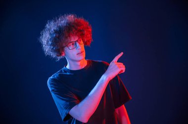 Parmakla göstererek sağı gösteriyor. Kıvırcık saçlı genç adam kapalı alanda neon ışıkla aydınlatılır..