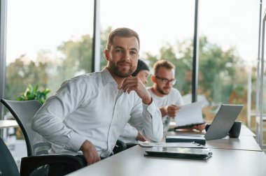 Adam kameraya bakıyor. Modern ofiste resmi kıyafetli üç kişi birlikte çalışıyor..
