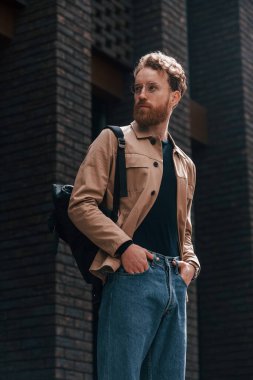 Sakallı, haki renkli ceketli ve kot pantolonlu şık bir adam binanın yanında..