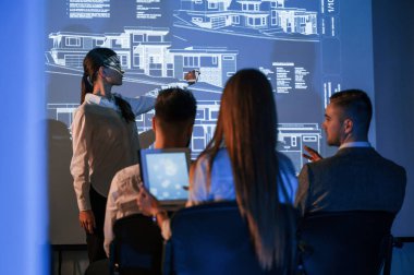 İşçilerle konuşan kadın lider, borsa şirketinin projektörüyle ilgili planı gösteriyor..