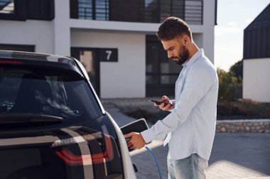 Aracı şarj etmek ve telefon kullanmak. Genç şık adam, gündüz vakti elektrikli arabayla geziyor..